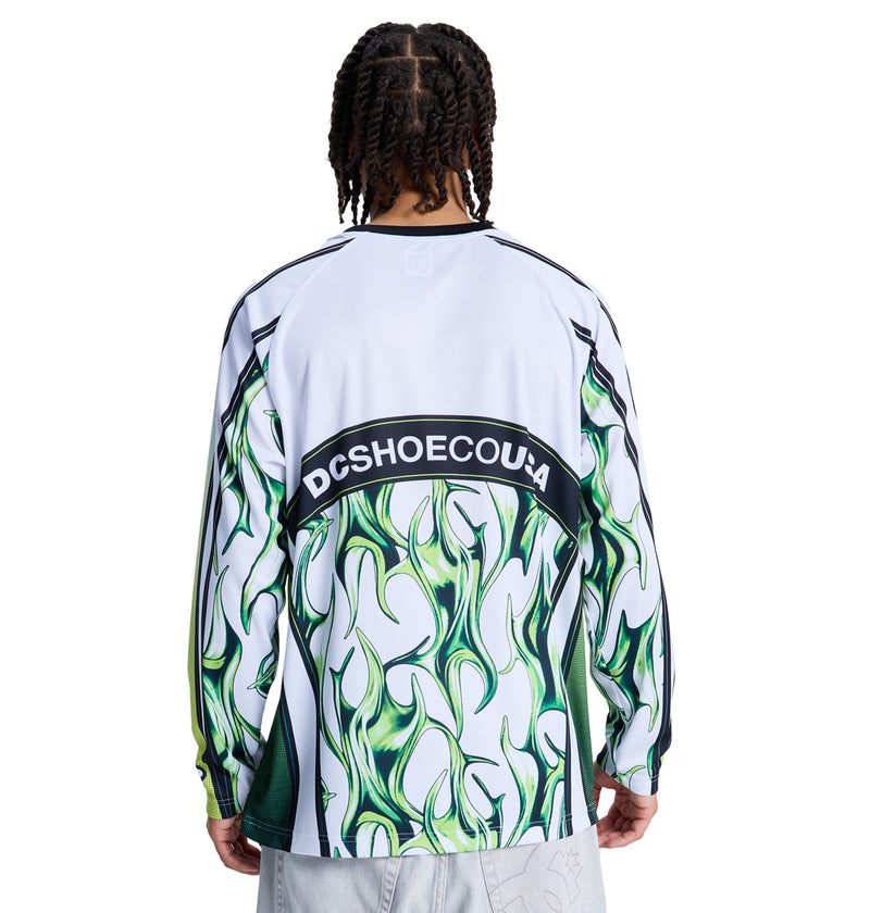 Person with braided hair wears the Reebok Sylem 2 Ls, a long-sleeve white shirt featuring green flame-like patterns and bold DCSHOECOUSA lettering across the back, standing against a plain background.