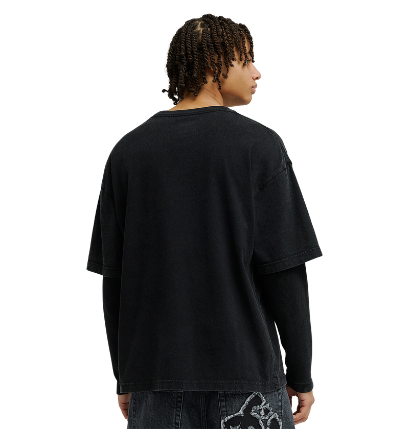 A person with short twists embodies streetwear style, wearing a loose black T-shirt layered over the DC Shoes Men’s DC Twofer Long Sleeve tee and black jeans with a white graphic, standing and facing away.