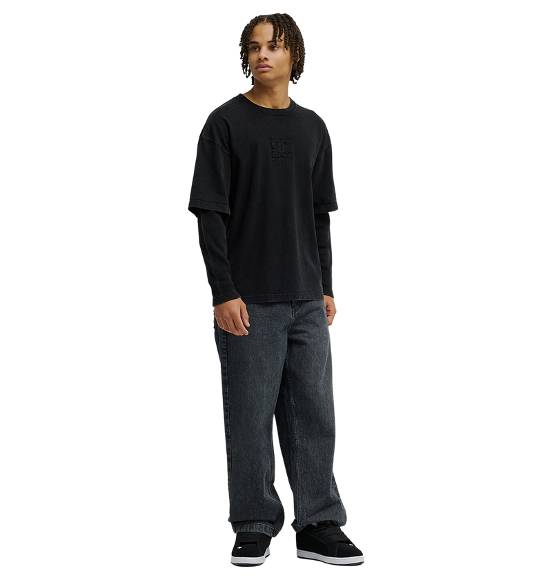 A person with medium-length curly hair wears the DC Shoes Men’s DC Twofer Long Sleeve under a black T-shirt, paired with loose dark jeans and black sneakers, embracing a streetwear style while standing against a plain white background.