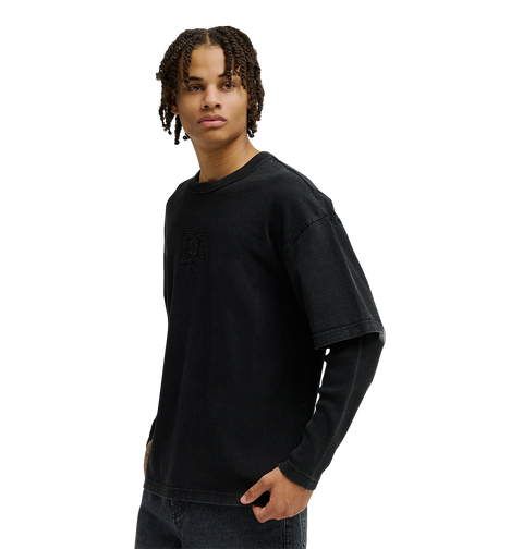 A person with short, curly hair stands against a plain white background, looking to the side, wearing a loose black T-shirt layered over the Men’s DC Twofer Long Sleeve by DC Shoes and paired with black jeans.