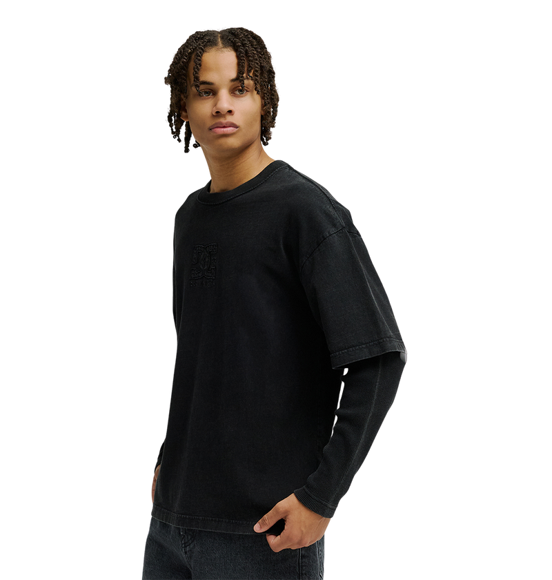 A person with short, curly hair stands against a plain white background, looking to the side, wearing a loose black T-shirt layered over the Men’s DC Twofer Long Sleeve by DC Shoes and paired with black jeans.