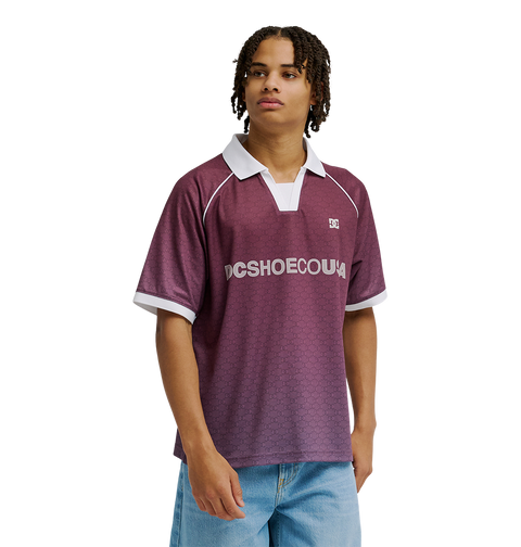 A young man models the Men's DC Soccer Top by DC Shoes, featuring maroon fabric with white trim, paired with light blue jeans against a plain white background for an athletic, casual look.