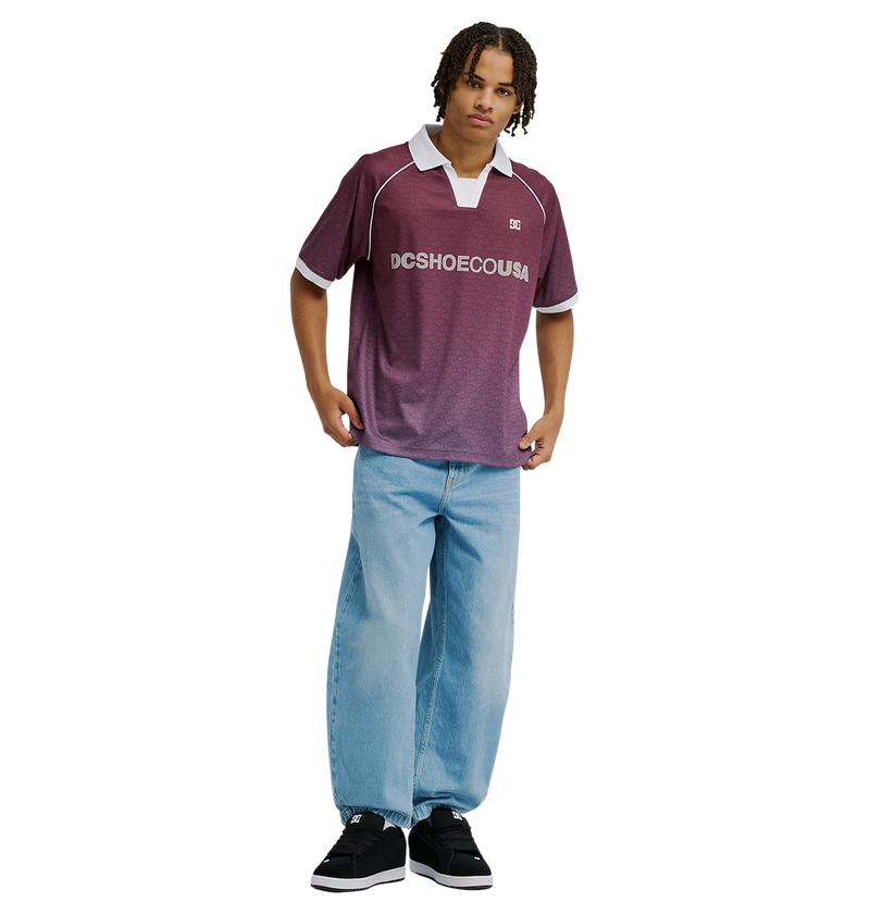 A person stands against a plain white background wearing the Men's DC Soccer Top by DC Shoes in maroon, paired with light blue baggy jeans and black sneakers.