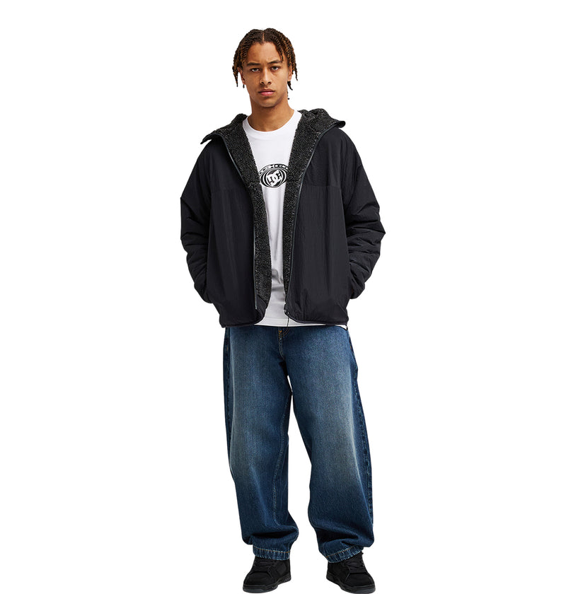A person stands facing forward, wearing the DC Shoes Coldstag Sherpa Reversible jacket—a black hooded fleece—paired with a white printed t-shirt, loose blue jeans, and black shoes. Their hands are tucked into their jacket pockets, exuding cold-weather comfort against a plain white background.