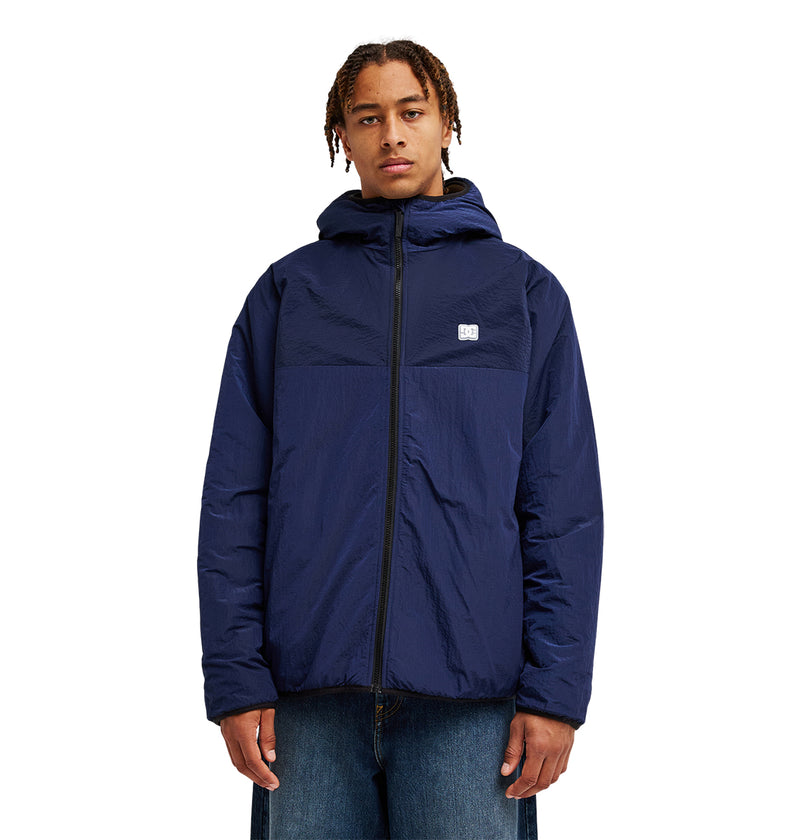 A person wearing the DC Shoes Coldstag Sherpa Reversible—a navy blue fleece jacket featuring a small white logo on the chest—stands facing forward against a plain white background, embodying cold-weather comfort.