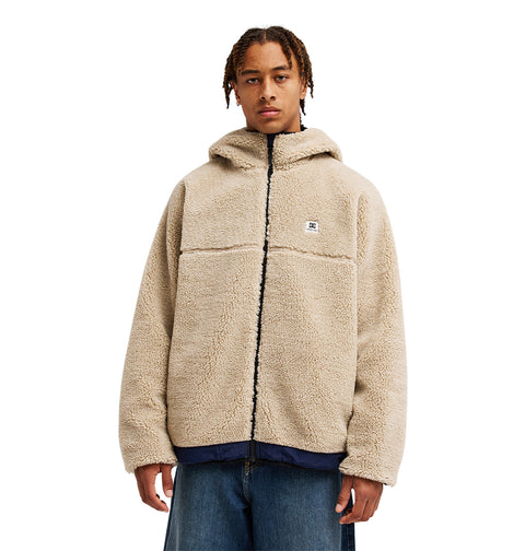 A person is wearing the DC Shoes Coldstag Sherpa Reversible jacket—a beige, hooded fleece with a small chest logo—paired with blue jeans, standing facing forward against a white background to highlight the cozy comfort of its warm fleece construction.