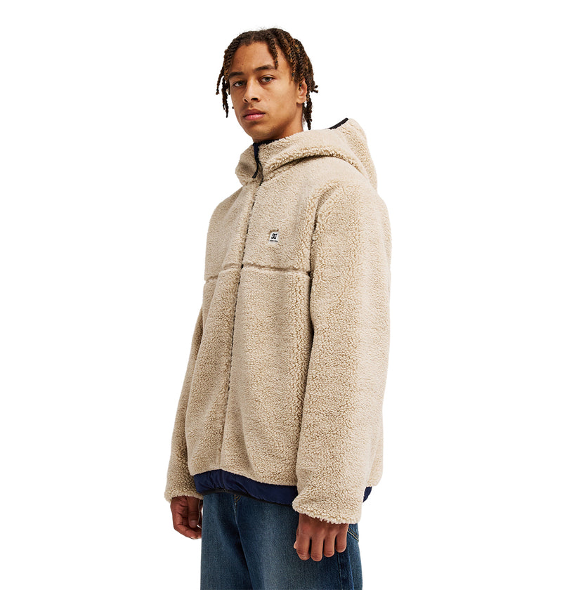 A person wearing the DC Shoes Coldstag Sherpa Reversible jacket in beige, featuring a cozy fleece construction, zip front, and hood, stands facing forward against a plain white background with blue jeans, highlighting the jacket’s exceptional cold-weather comfort.