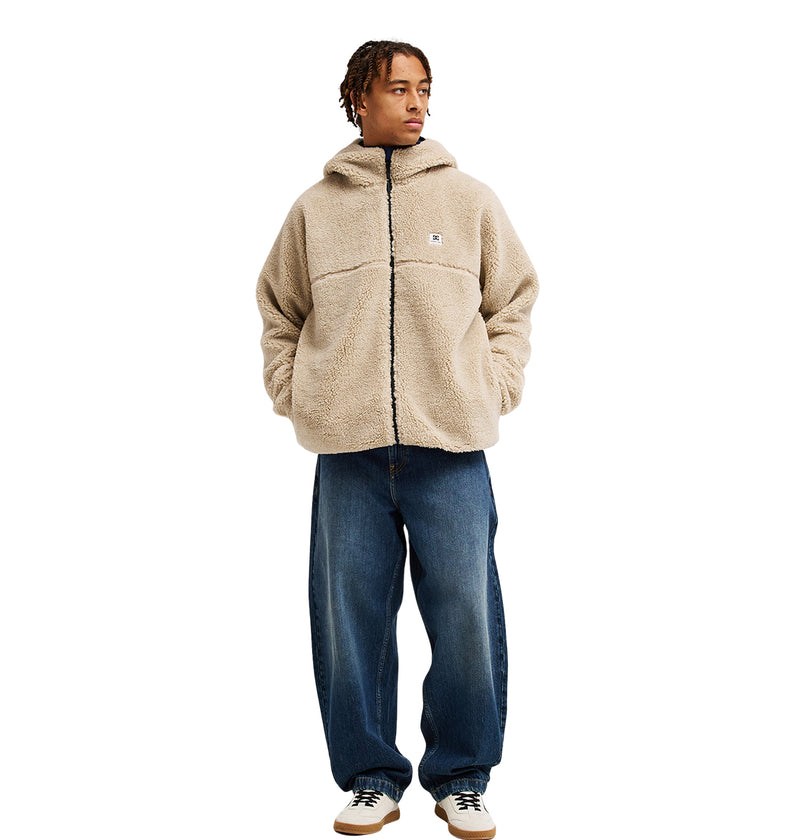 A person stands facing forward with their hands in their pockets against a white background, wearing the DC Shoes Coldstag Sherpa Reversible—a beige zip-up jacket with a hood, crafted from cozy fleece for cold-weather comfort—paired with wide blue jeans and sneakers.
