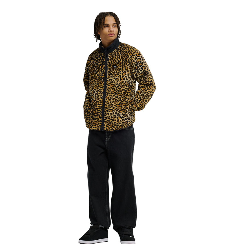 A person stands against a plain white background wearing the DC Shoes Lynx Fleece jacket in yellow and black leopard print, providing soft fleece warmth and cold-weather comfort, paired with black pants and black shoes.