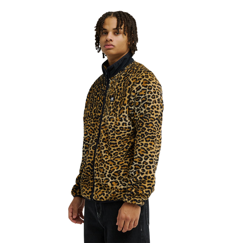 A person with short dreadlocks stands facing slightly to the side against a white background, wearing a leopard print DC Shoes Lynx Fleece zip-up jacket and black pants, highlighting the cozy cold-weather comfort and soft fleece style of the DC Shoes Lynx Fleece.