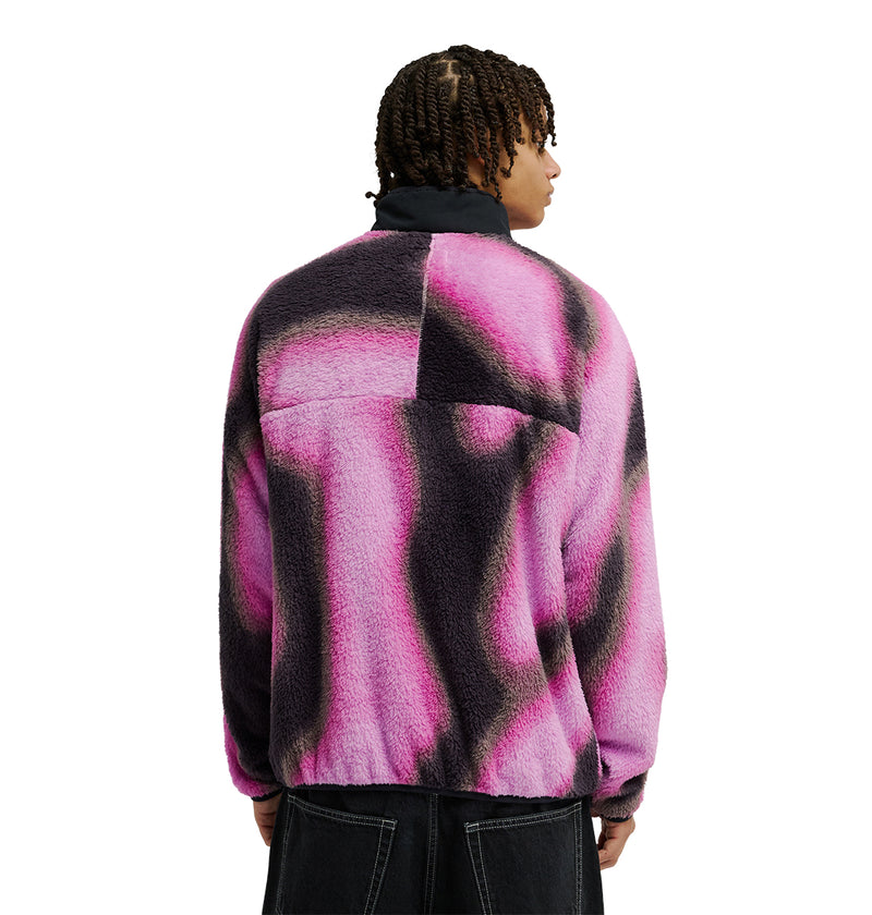 Wearing the DC Shoes Lynx Fleece, a fuzzy jacket in pink, purple, and black abstract patterns designed for cold-weather comfort with practical pockets, a person with short twists is seen from the back against a plain white background.