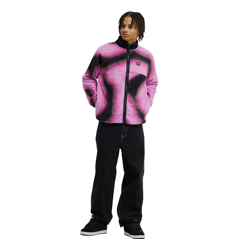 Wearing the DC Shoes Lynx Fleece in a pink and black pattern, this person stands with hands in practical pockets, paired with loose black pants and black shoes, against a white background.