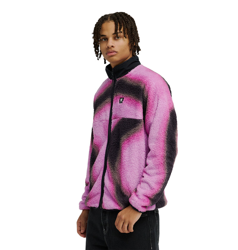 A person with braided hair wears the DC Shoes Lynx Fleece—a soft, fuzzy pink and black zip-up jacket with a black collar—paired with black pants, standing against a plain white background.