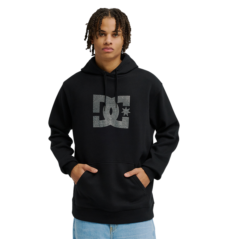A person wearing the Men’s DC Strass Star Hoodie by DC Shoes, featuring a large rhinestone DC star logo on the front, stands with hands in the pocket. The black hoodie includes a drawstring hood for cozy fleece warmth, and is paired with light blue jeans.