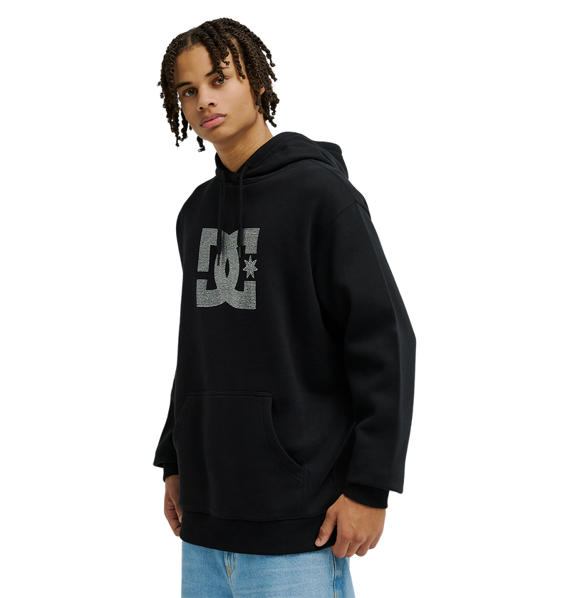 A person with short, curly hair models the Men’s DC Strass Star Hoodie by DC Shoes in black, paired with light blue jeans. Standing against a plain white background and gazing slightly to the side, they showcase the hoodie’s iconic DC logo and its blend of classic fleece warmth with modern style.