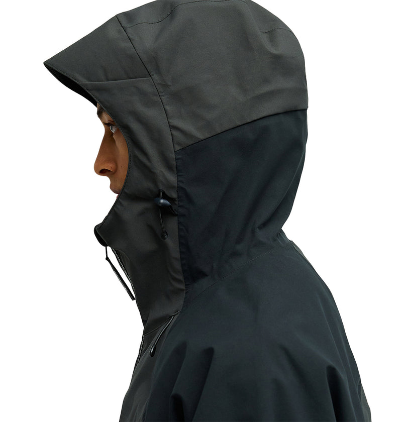 A person in profile view, facing left, wears the DC Shoes Storm Divison 3L Jacket 30K—a dark waterproof jacket with the hood up. The jacket features a drawstring and offers breathable protection, set against a plain white background.