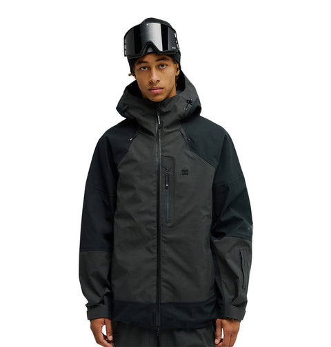 A person stands facing forward against a plain white background, wearing the DC Shoes Storm Division 3L Jacket 30K, a dark, breathable protective snow jacket, with ski goggles resting on their head.