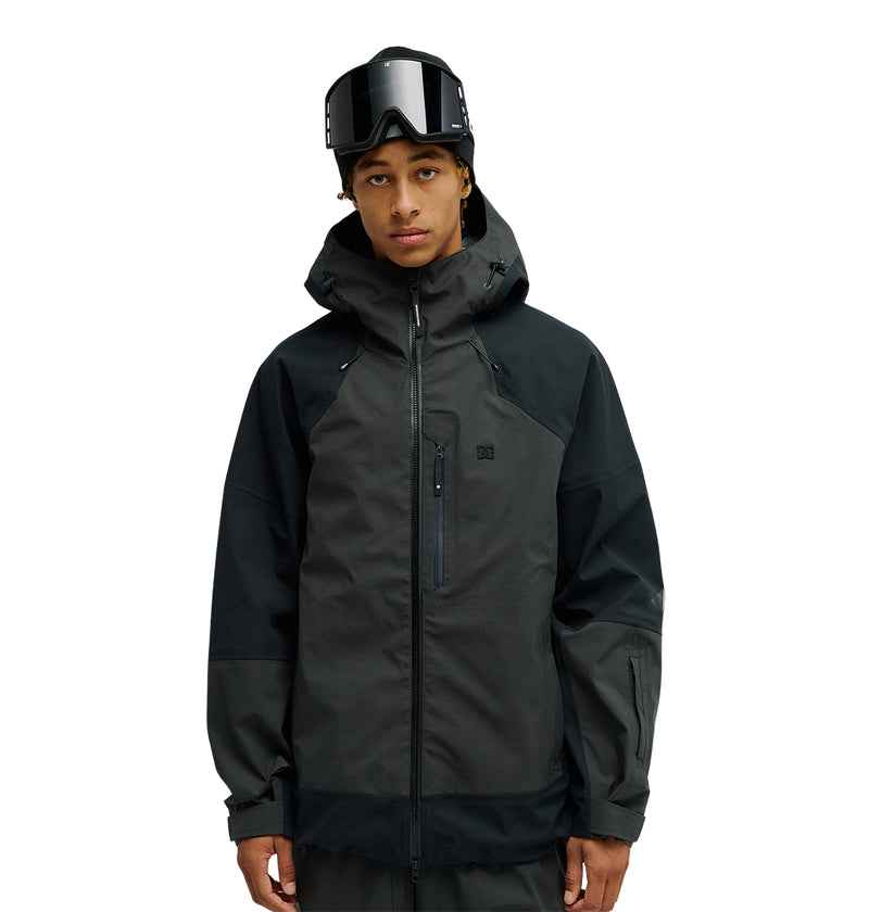 A person stands facing forward against a plain white background, wearing the DC Shoes Storm Division 3L Jacket 30K, a dark, breathable protective snow jacket, with ski goggles resting on their head.