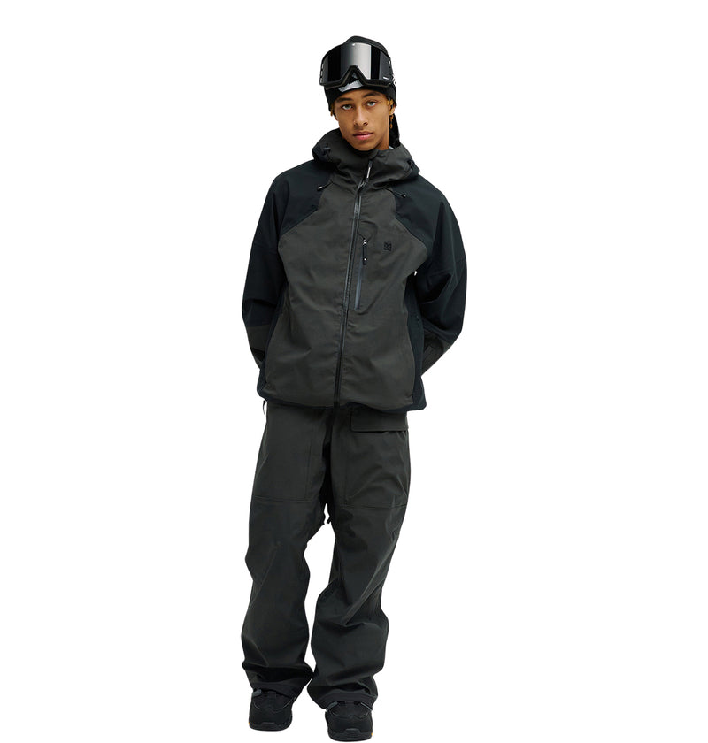 A person wearing the DC Shoes Storm Division 3L Jacket 30K stands against a white background with ski pants, boots, and goggles, hands behind their back and looking slightly to the side.