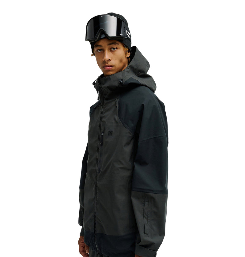 A person wearing the DC Shoes Storm Division 3L Jacket 30K in a dark color, paired with ski goggles, stands facing slightly to the side against a plain white background.