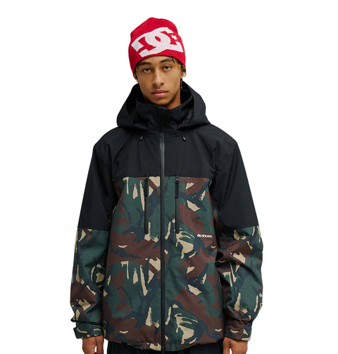 A person wearing a red DC Shoes beanie and a black and camouflage DC Shoes Edge Jacket 20K with a front zipper, featuring advanced waterproofing for superior snow performance, stands against a plain white background.