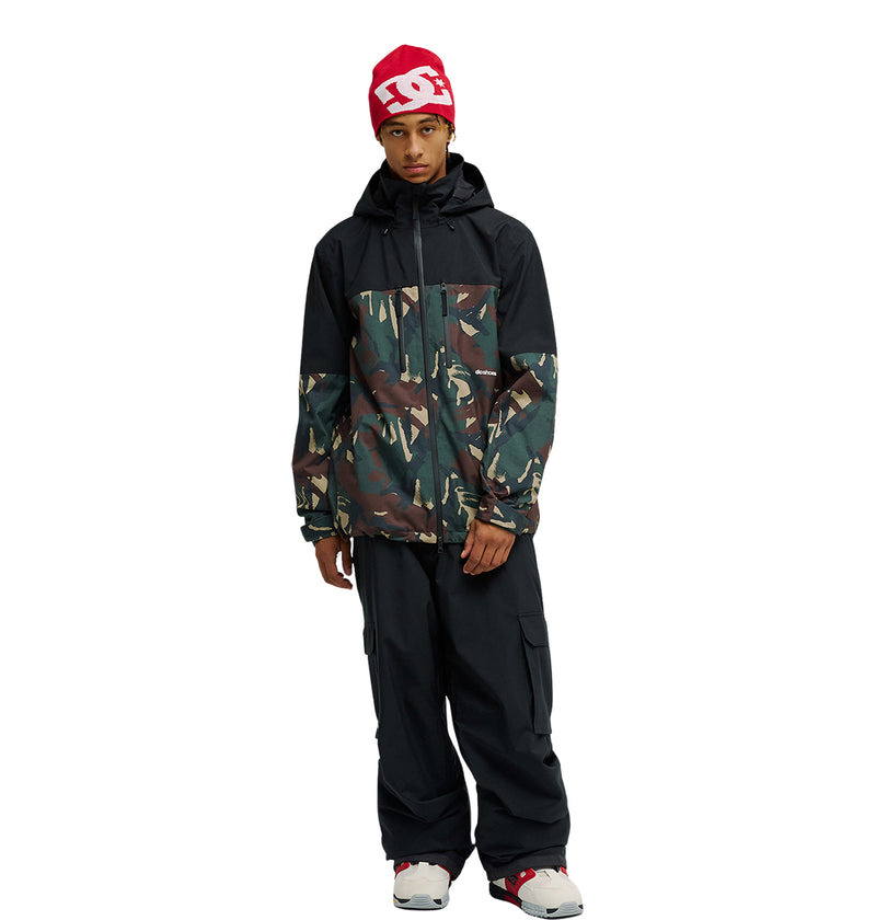 A person wears a red beanie, a black DC Shoes Edge Jacket 20K featuring a camouflage pattern engineered for exceptional snow performance, black cargo pants, and white sneakers with red accents, standing against a plain white background.
