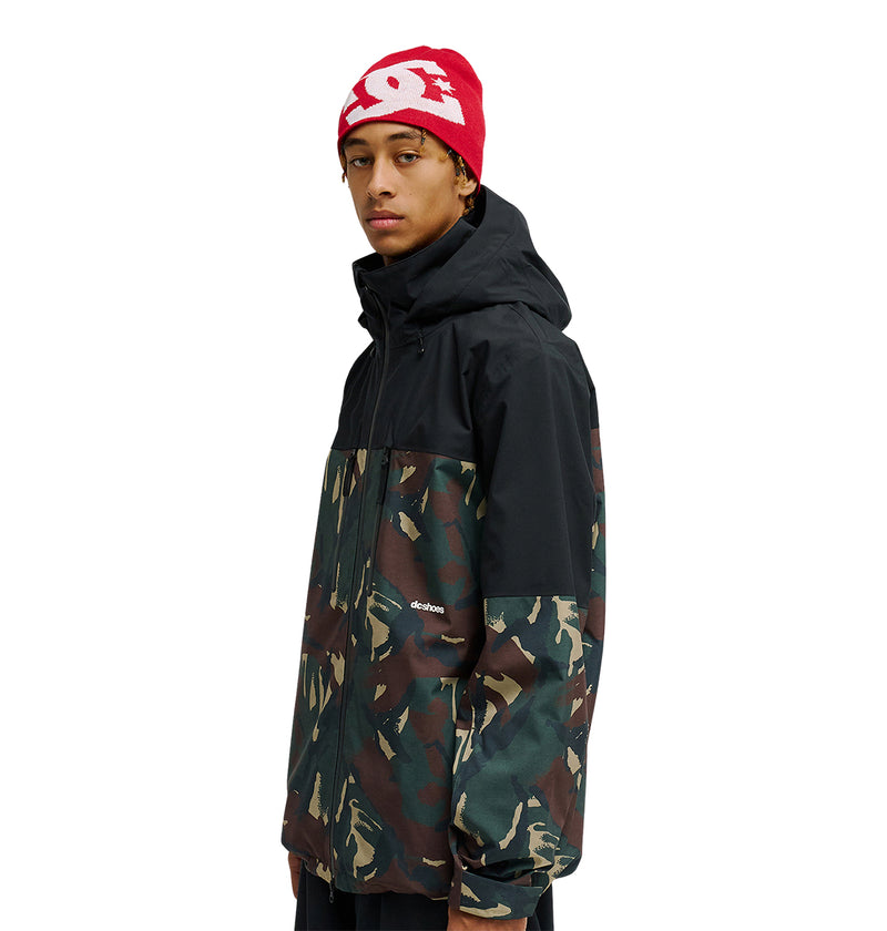 A person wearing the DC Shoes Edge Jacket 20K in black and camouflage, featuring a hood and designed for snow performance with enhanced waterproofing, stands facing sideways against a white background. They are also wearing a red beanie with a white logo.
