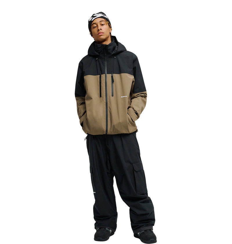 A person stands against a plain white background wearing the DC Shoes Edge Jacket 20K—a two-tone black and brown waterproof jacket—paired with black cargo pants, black shoes, and a black and white beanie.