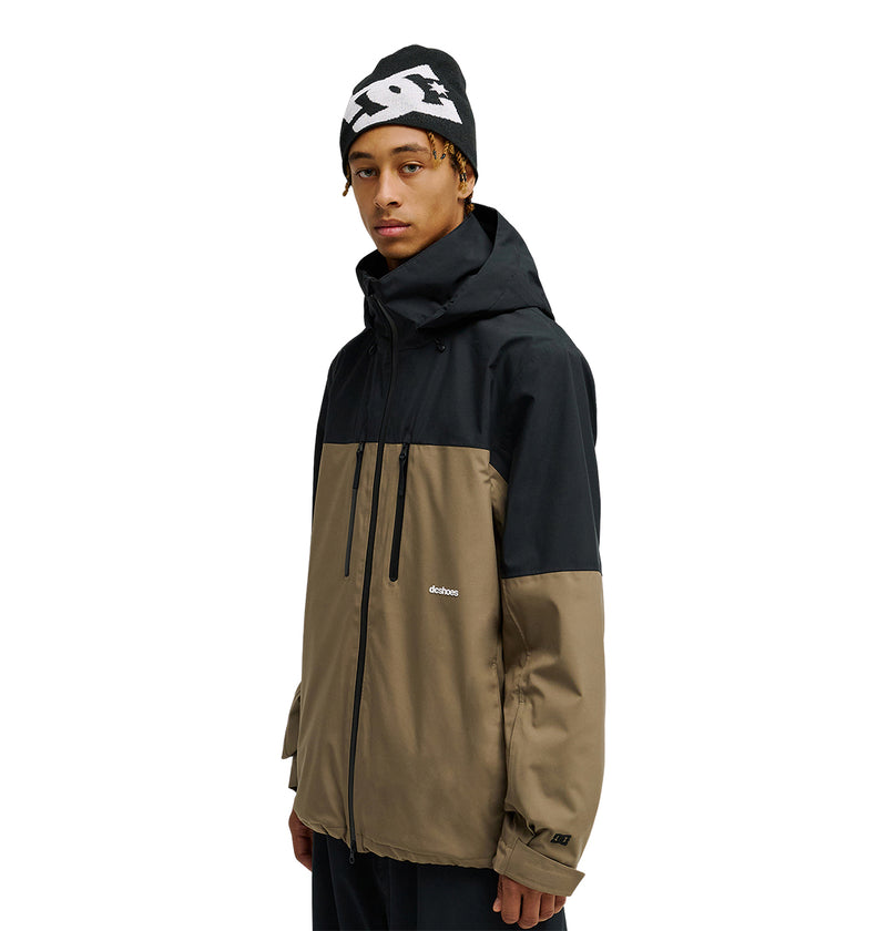 A person wearing the DC Shoes Edge Jacket 20K in black and tan, featuring “dcshoes” on the chest, along with a black beanie with a white pattern, standing against a white background.