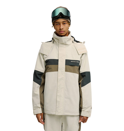 A person stands facing the camera against a plain white background, wearing the DC Shoes Mountain Lab Jacket 20K in a beige and olive color-block design with matching pants, featuring advanced waterproofing for snow performance, and has ski goggles resting on their head.