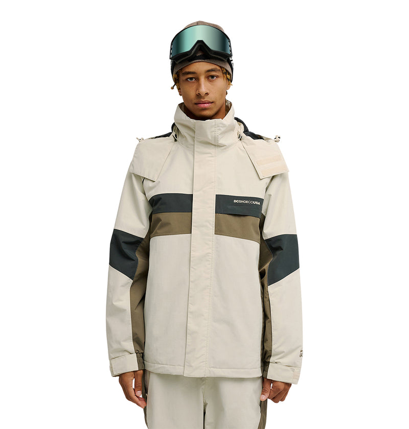 A person stands facing the camera against a plain white background, wearing the DC Shoes Mountain Lab Jacket 20K in a beige and olive color-block design with matching pants, featuring advanced waterproofing for snow performance, and has ski goggles resting on their head.