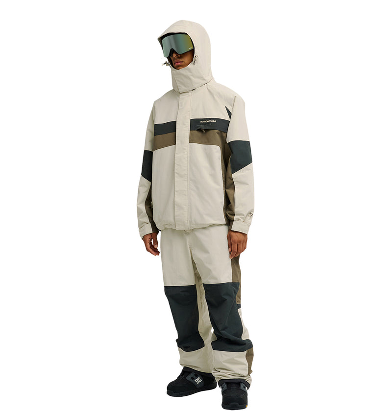 A person is wearing the DC Shoes Mountain Lab Jacket 20K, a beige and gray hooded winter jacket designed for breathable protection and enhanced snow performance, paired with matching pants, black boots, and large reflective ski goggles, standing against a white background.