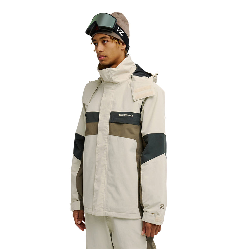 A person wearing the DC Shoes Mountain Lab Jacket 20K in beige and olive green, featuring breathable protection, along with matching pants, a brown beanie, and ski goggles resting on their head, stands against a white background.