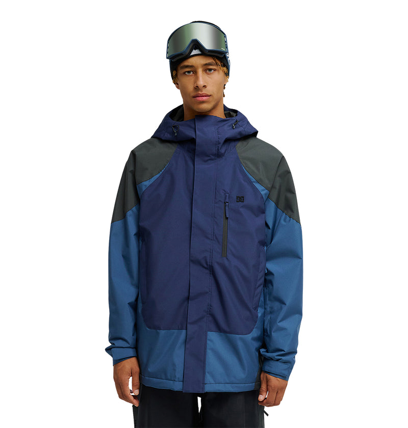 A person wearing the DC Shoes Coldsmoke Jacket 10K in blue and navy, paired with ski goggles, stands facing forward against a white background, highlighting the jacket’s superior snow performance and breathable protection.