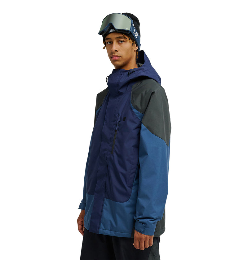 A person wearing the DC Shoes Coldsmoke Jacket 10K in blue and dark gray, with ski goggles on their head, standing against a plain white background.
