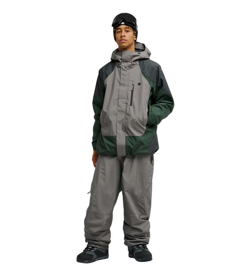A person wearing the DC Shoes Coldsmoke Jacket 10K in gray and green—with advanced waterproofing and breathable protection—along with gray snow pants, black boots, and black ski goggles stands against a white background.