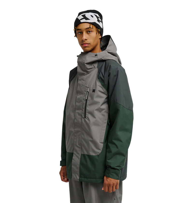A person stands facing slightly left against a white background, wearing the DC Shoes Coldsmoke Jacket 10K in dark green and gray with a hood, along with a black beanie featuring white text.