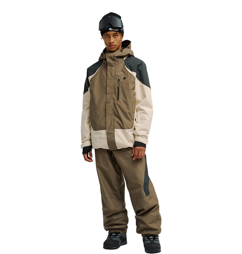 A person wearing the DC Shoes Coldsmoke Jacket 10K in brown and beige—designed for excellent snow performance—paired with matching ski pants, black gloves, black boots, and a black ski helmet with goggles, standing against a plain white background.