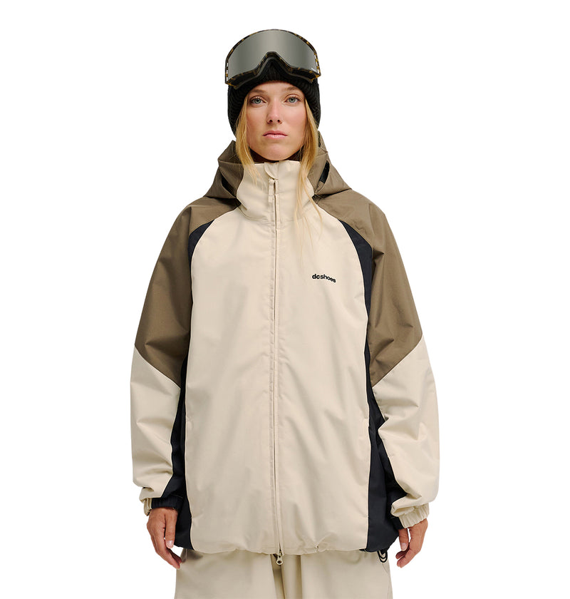 A woman stands facing forward against a white background, wearing the DC Shoes Trackstar Jacket 10K in beige and brown for breathable protection, paired with tan pants, black gloves, and ski goggles on her head.