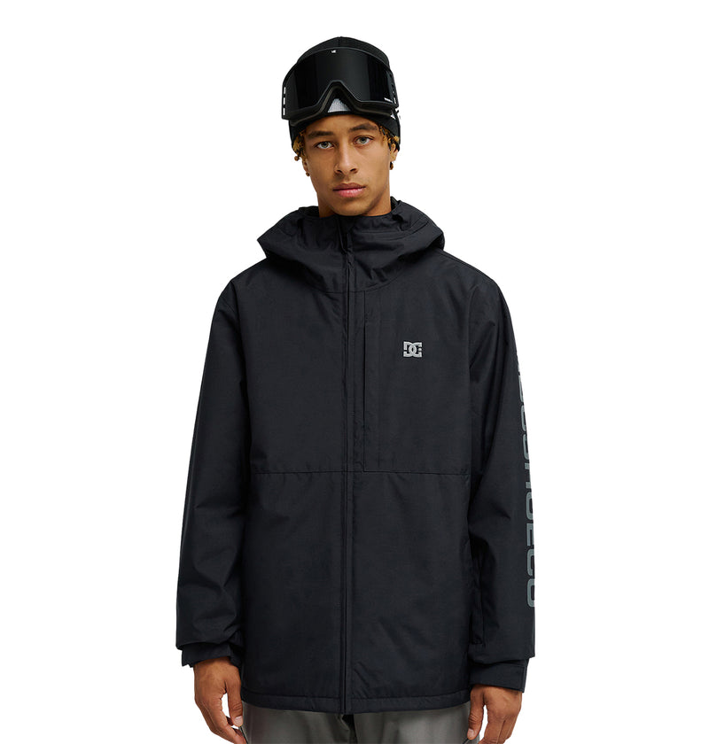 A person wearing a black DC Shoes Basis Jacket 10K—an insulated, breathable, and waterproof snow jacket featuring a small logo—paired with gray pants and ski goggles on their head, standing against a white background.