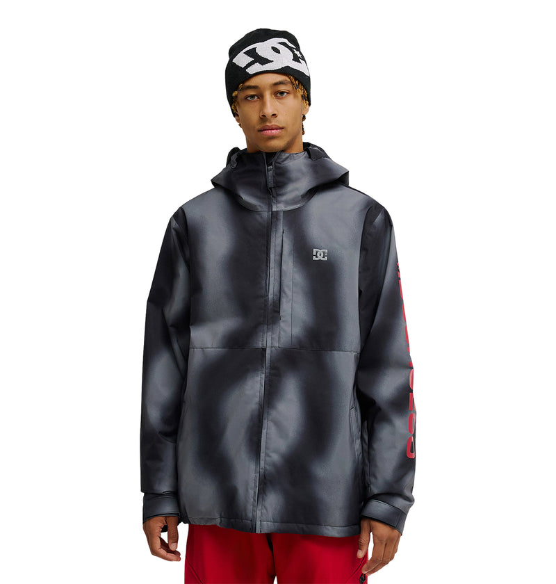 A person wearing the DC Shoes Basis Jacket 10K, an insulated black and gray jacket with a logo on the chest, along with red pants and a black beanie featuring a white pattern, standing against a white background.