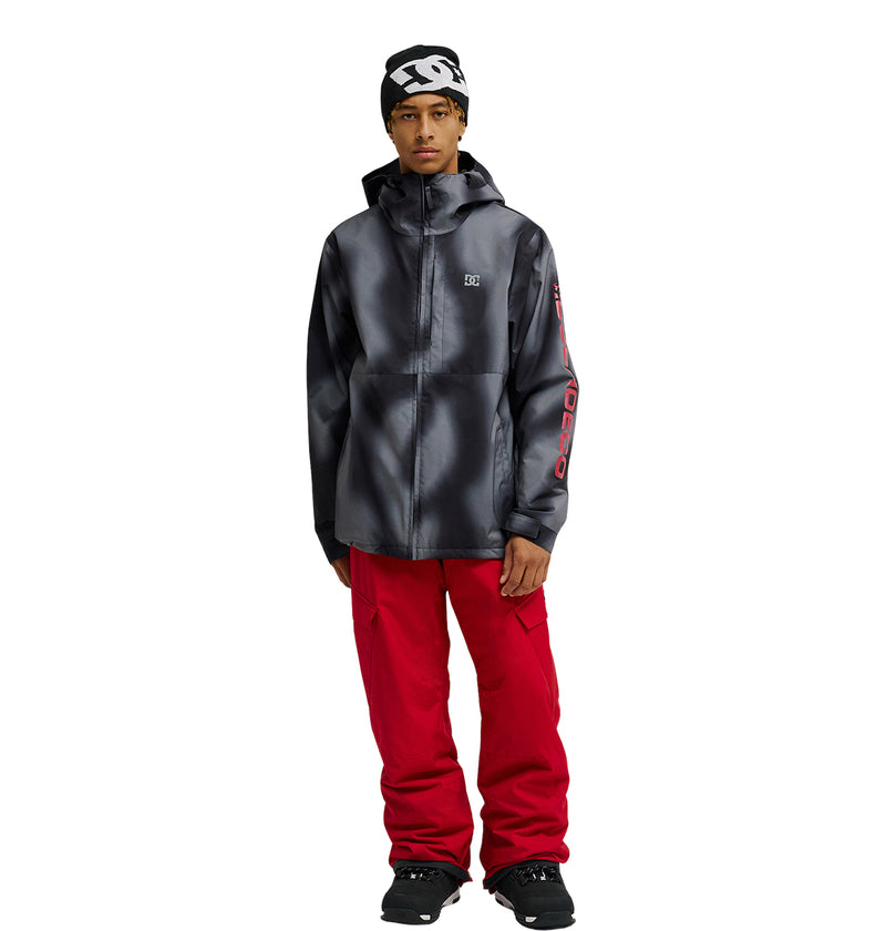 A person wearing the DC Shoes Basis Jacket 10K—an insulated black and gray hooded jacket—paired with red pants, black shoes, and a black beanie with white patterns, stands against a white background.