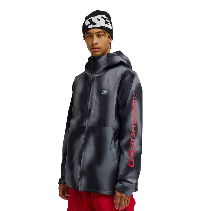A person wearing a black DC Shoes Basis Jacket 10K with a hood, featuring a red DC SHOES logo on the sleeve, red pants, and a black beanie with a white DC Shoes logo, standing against a white background.