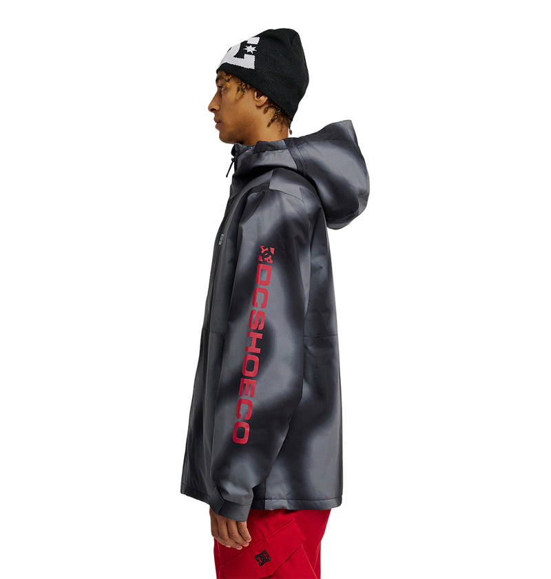 A person wearing a black beanie, bright red pants, and the DC Shoes Basis Jacket 10K—a dark gray insulated jacket featuring “DCSHOECO” in red on the sleeve—stands in profile against a white background.