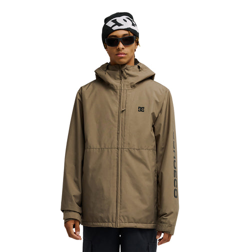 A person stands against a white background wearing the DC Shoes Basis Jacket 10K—an insulated, tan hooded snow jacket featuring the DC logo, black beanie, sunglasses, and black pants. The left sleeve displays “DC SHOES CO” print. Offering 10K waterproofing and breathable protection, this jacket is designed for all your winter adventures.