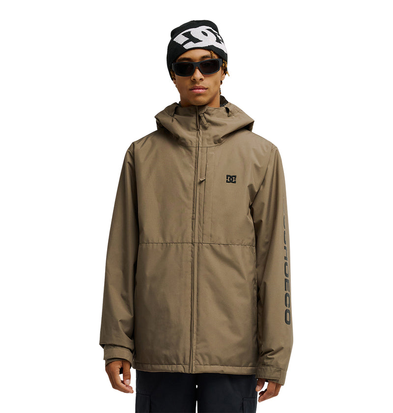 A person stands against a white background wearing the DC Shoes Basis Jacket 10K—an insulated, tan hooded snow jacket featuring the DC logo, black beanie, sunglasses, and black pants. The left sleeve displays “DC SHOES CO” print. Offering 10K waterproofing and breathable protection, this jacket is designed for all your winter adventures.