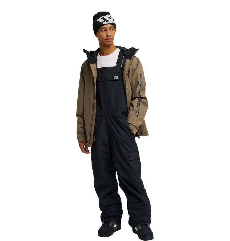 A person wearing a black beanie, a white T-shirt, and black snow bib pants stands with hands in their pockets while layered in the DC Shoes Basis Jacket 10K against a white background.