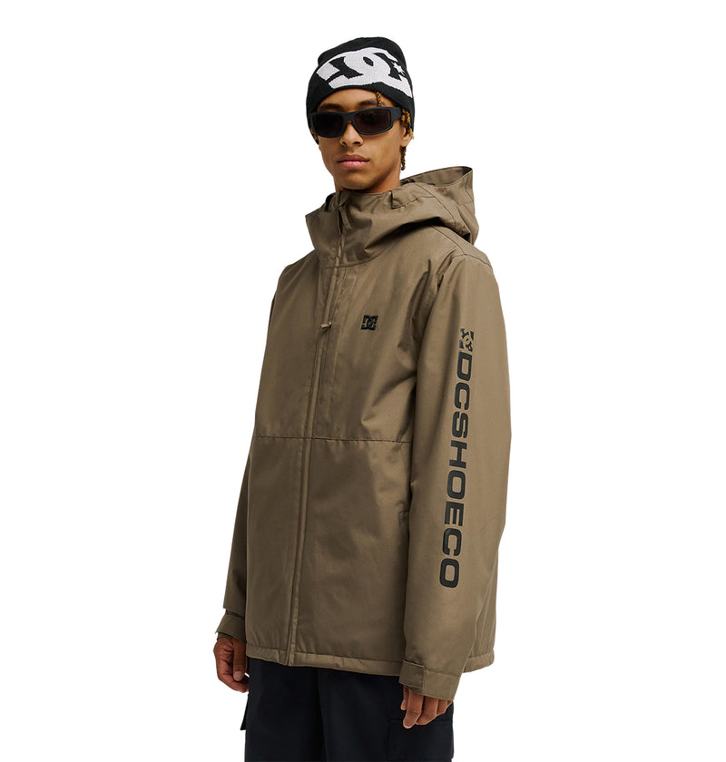 A person wearing the DC Shoes Basis Jacket 10K in tan with “DCSHOECO” on the sleeve, paired with a black beanie featuring a white design and sunglasses, standing against a plain white background.