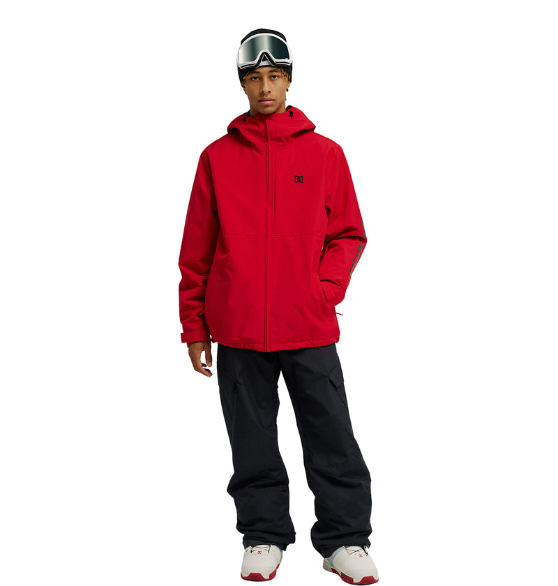 A person wears the DC Shoes Basis Jacket 10K in red, paired with black snow pants, white sneakers, and ski goggles, standing facing forward against a white background.