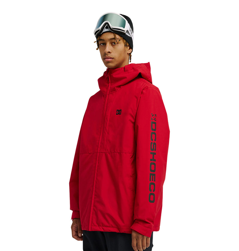 A person wearing the DC Shoes Basis Jacket 10K in red, featuring DCSHOECO branding on the sleeve, paired with black pants and ski goggles resting on their head, stands against a white background.